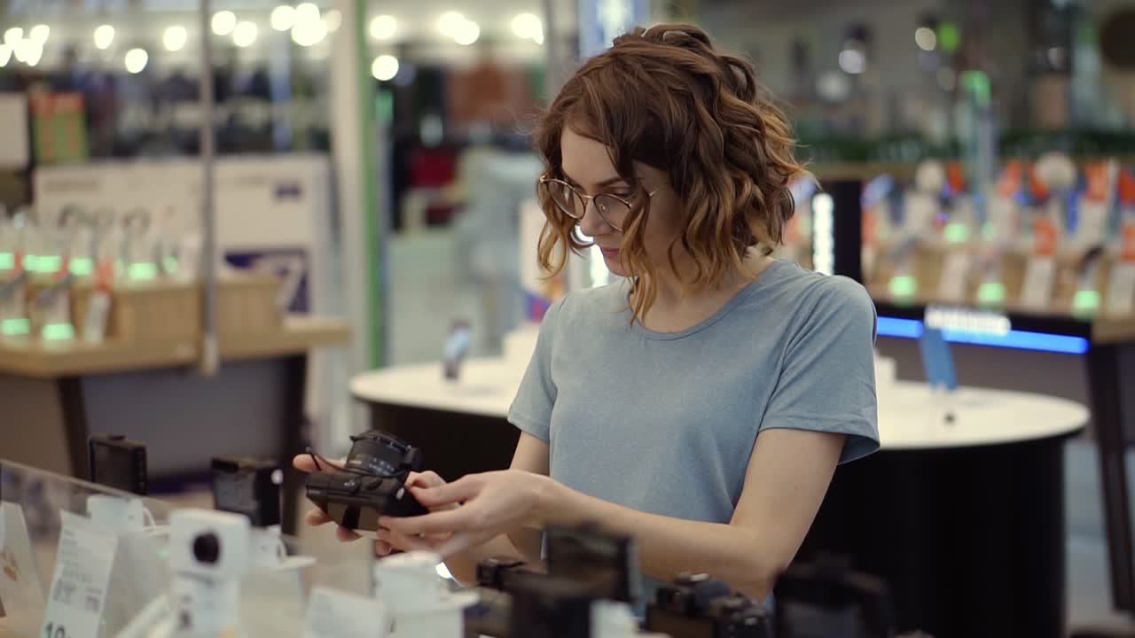 Young curly woman in glasses shopping for a new photocamera in the electronics store. Trying to decide on the best model. Have Doubts. Checking one camera in hands