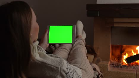 mujer acostada en el sofá frente a la chimenea sosteniendo una pantalla verde de tableta, viendo la pantalla clave de croma junto a la chiminea