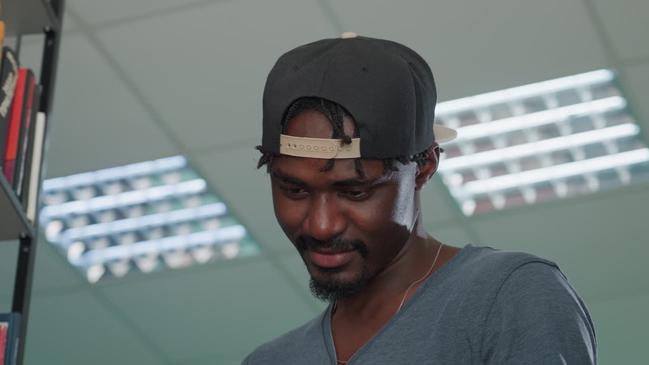 Close up of young man wearing backward cap and casual shirt smiling subtly while reading book in library under bright fluorescent lights, showing relaxed and focused expression