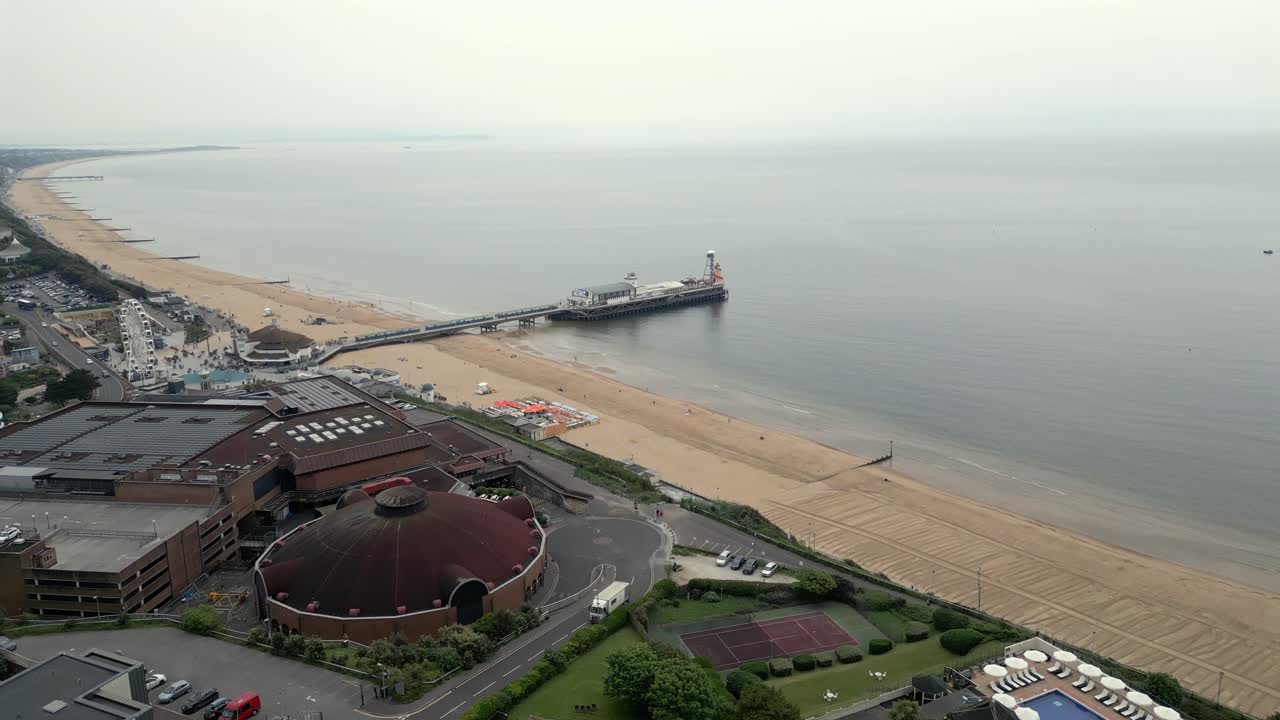 A smooth aerial dolly shot of Bournemouth beach, iconic pier, and surrounding coastal scenery under an overcast sky.