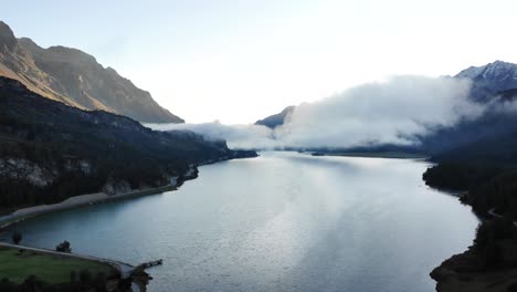 Un-Sobrevuelo-Aéreo-Del-Amanecer-Sobre-El-Lago-Sils-En-Maloja,-Suiza,-Con-Vistas-A-Los-Picos-De-Engadin-Y-Nubes-Sobre-El-Agua