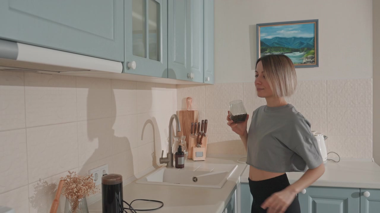 Donna bianca in cucina amante della musica tocca l'altoparlante mentre si allunga verso il pensile superiore e prepara il caffè, mobili color pastello, piccolo altoparlante Bluetooth nero sul bancone, tazza di ceramica, fiori secchi, incorniciato