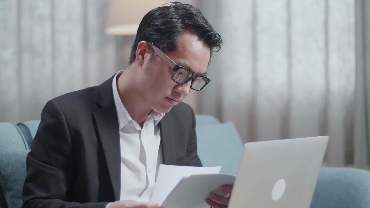 un hombre de negocios asiático en chaqueta y pantalones cortos chequeando los documentos mientras trabaja en una computadora portátil en casa