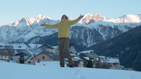 joven caucásico con chaqueta amarilla y gorro que se detiene para ver la vista del pequeño pueblo de montaña y la iglesia y levanta las manos en guarda, suiza