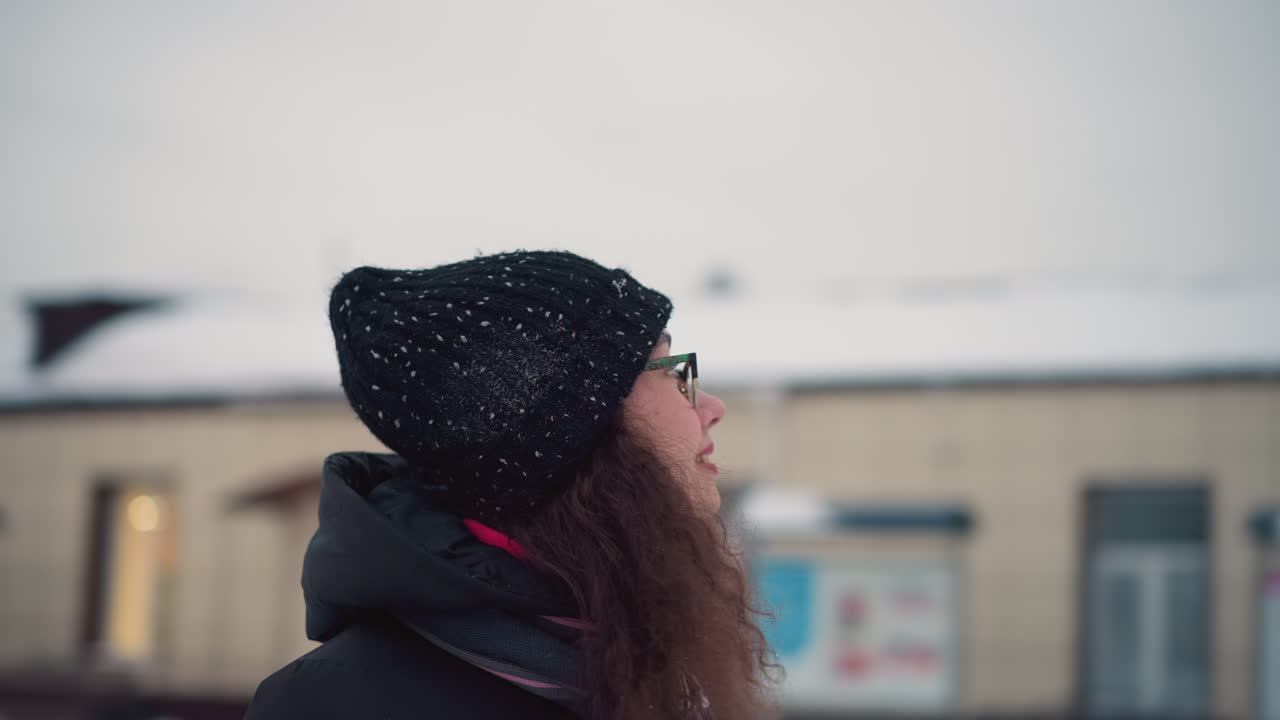 Skater with curly hair wearing black winter hat, glasses, and striped scarf smiles while looking upward in snowy outdoor setting, soft winter lights glow in background creating cozy seasonal atmosphere