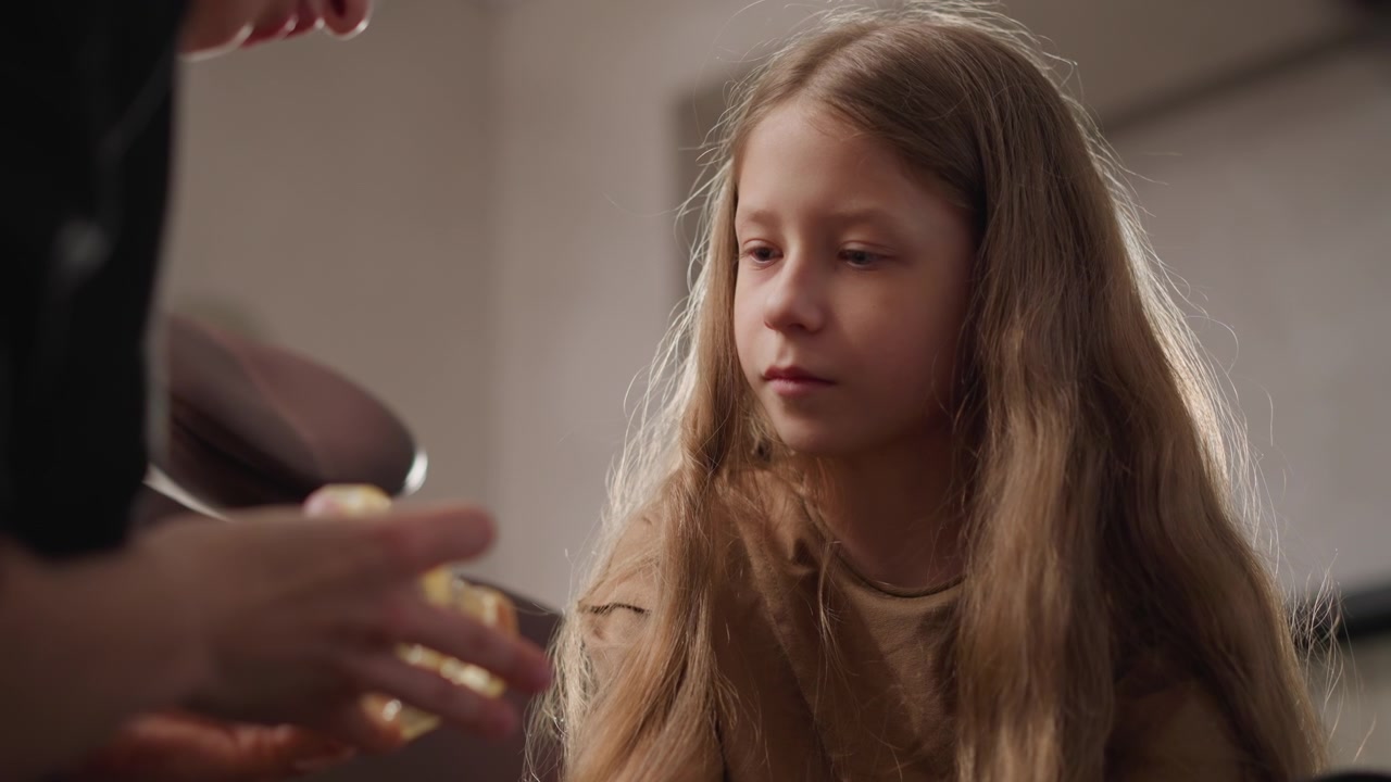 Close up of young girl receiving hair treatment from adult in soft lighting. child sits still as another girl gently combs and styles her long blonde hair with hands visible performing