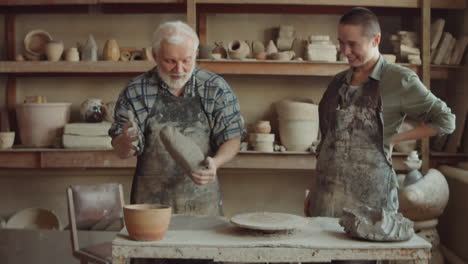 senior potter kneading clay and teaching young woman in workshop