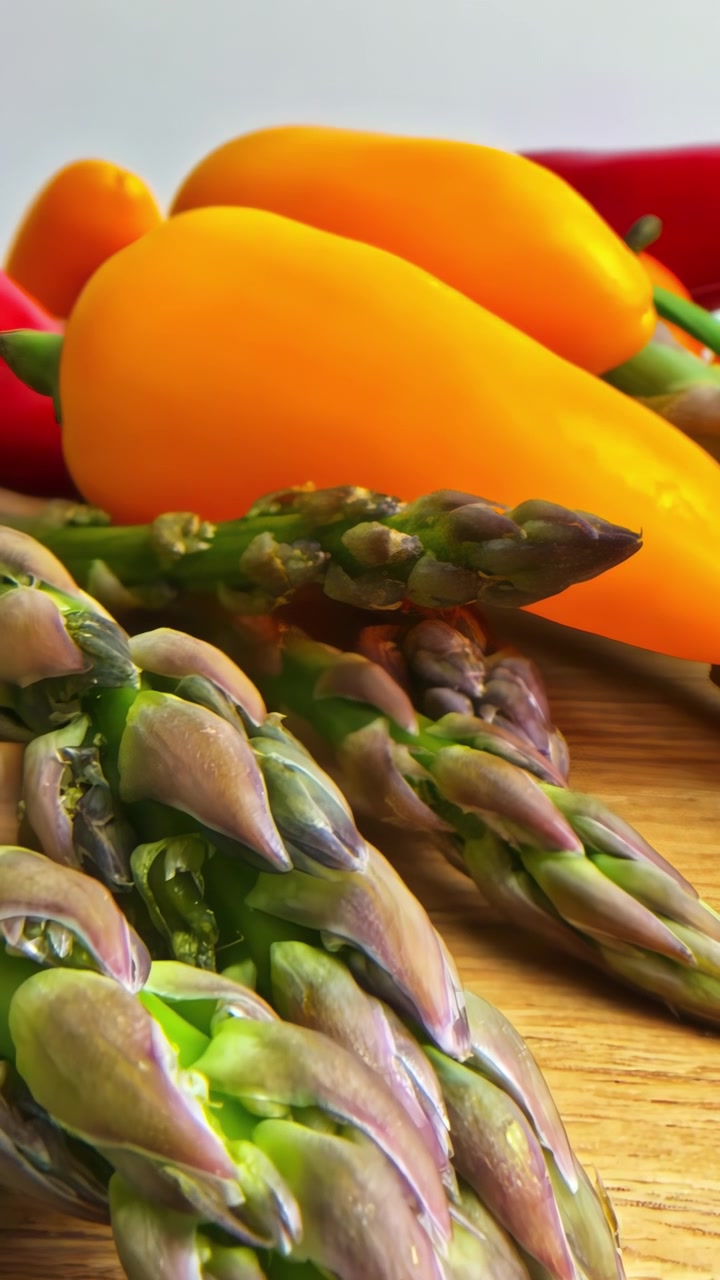 Pushing in over vibrant asparagus spears with purple tips resting on a warm wooden board, yellow and red peppers softly blurred behind Embodying the quiet abundance of fresh produce.