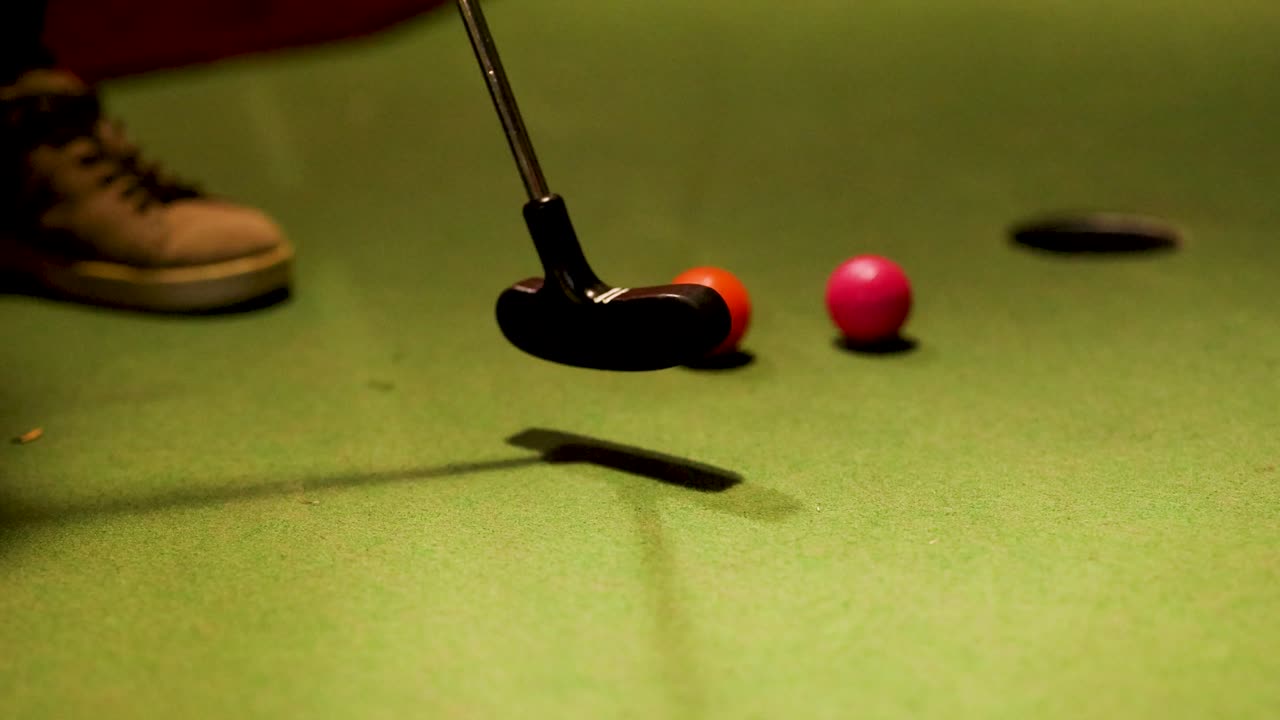 A mini golf putter hits two colorful balls on a green course under warm lighting