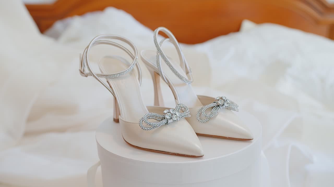Elegant white bridal heels with rhinestone bows on soft bed, indoor set romantic