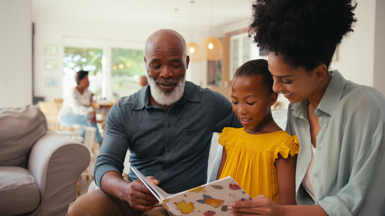 Großvater mit Enkelin und Mutter lesen zu Hause ein Buch, mit Mehrgenerationenfamilie im Hintergrund