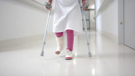 lowsection of african american girl in hospital gown walking using crutches in corridor, slow motion