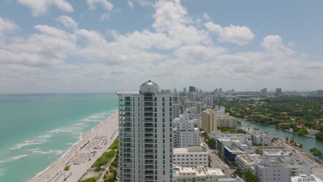 Modern-building-at-sea-coast.-Aerial-descending-shot-of-high-rise-apartment-building-near-sand-beach.-Miami,-USA