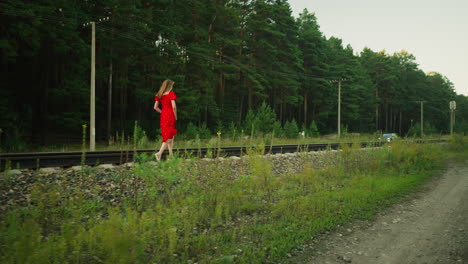young lady in red dress runs carefully across railway sleeper holding gown for balance, with parked car and electric poles in background, surrounded by gravel, grass, and dense green forest