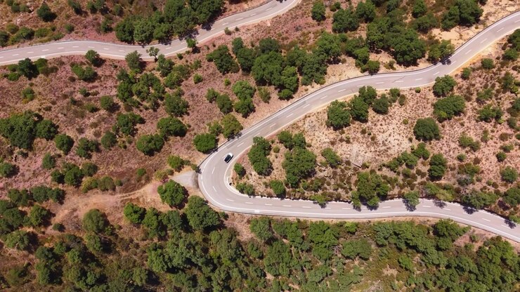 Vista dall'alto di una strada di montagna tortuosa che attraversa una macchia arida e alberi verdi, con una singola auto nera che percorre curve strette in una giornata di sole