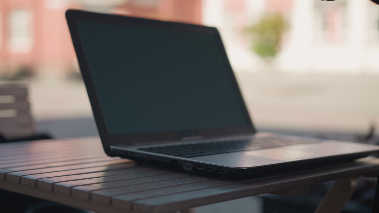 Close-up of person in black coat picking up coffee cup from table with laptop nearby, table setup includes coffee cup and open laptop with blurred background featuring buildings and greener