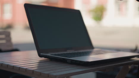 primer plano de una persona con una chaqueta negra recogiendo una taza de café de la mesa con una computadora portátil cerca, la configuración de la mesa incluye una tazón de café y una computadora portable abierta con un fondo borroso con edificios y más verde