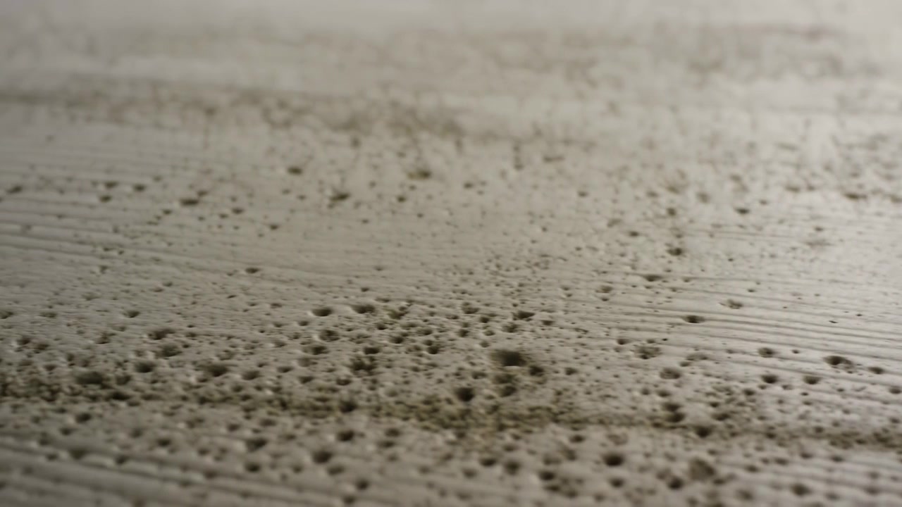 Close-up of a rough beige surface filling the frame, with the main area concentrated in the lower center and foreground. Fine grain, pits, and worn parallel