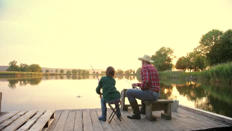 widok z tyłu nastoletniego chłopca siedzącego z dziadkiem na molo nad jeziorem, rozmawiającego i łowiącego razem ryby