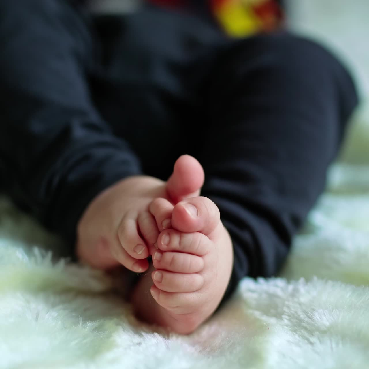 Cute baby feet rubbing each other. Child in dark clothes lies on the bed covered with white plaid. Blurred backdrop