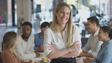 portrait of businesswoman with colleagues in background sitting around table in open plan office