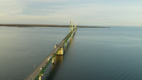 High-aerial-following-traffic-driving-over-long-suspension-bridge,-calm-water,-wide-shot