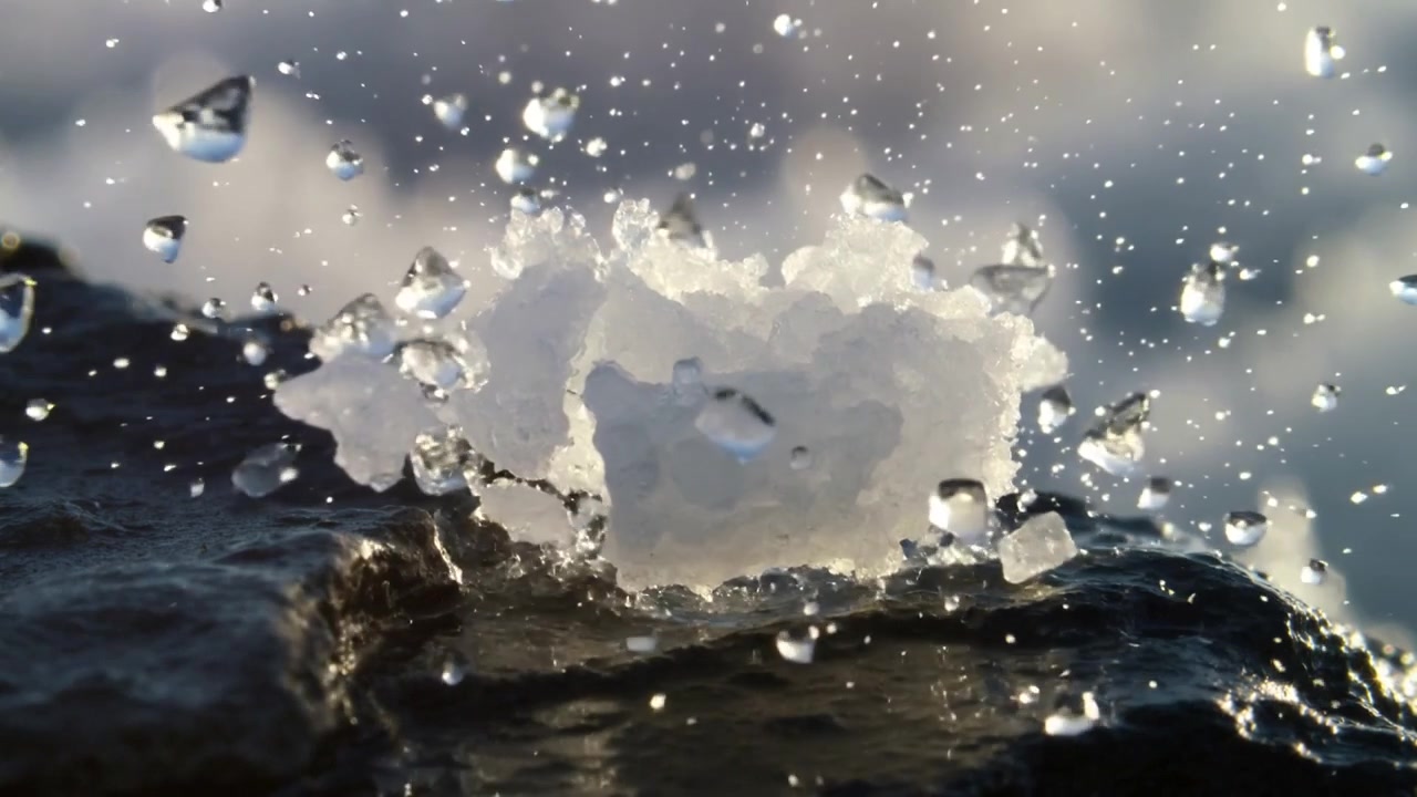Druppel raakt een klein klompje sneeuw en ijs op een donkere natte rots nabij de kustlijn, waarbij druppels worden weggeslingerd.