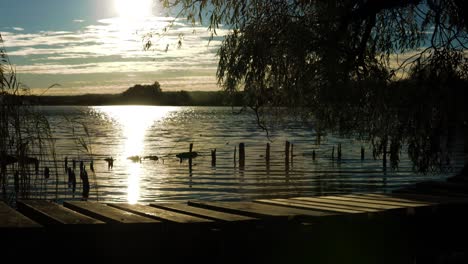 Eine-Dunstige-Sonnenuntergangsaufnahme-Eines-Mannes,-Der-Auf-Einer-Kleinen-Holzbrücke-über-Das-Wasser-Geht