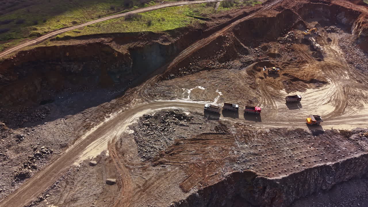 Workers move machinery and trucks around a granite quarry during daylight hours in a remote location with various rock formations visible