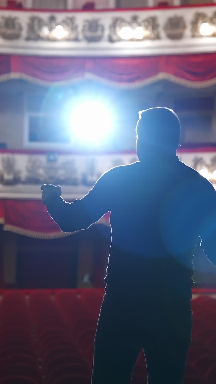 Announcer on a stage in theater. Bright round light on one person. Man in black suit performs to the auditorium. View from the back. Vertical video
