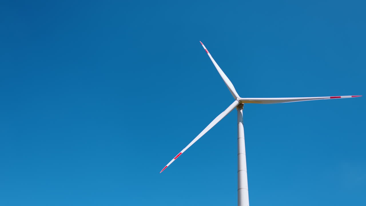 Tall wind turbine, blue sky. A large wind turbine rotates slowly, contrasting vividly with the clear blue sky above