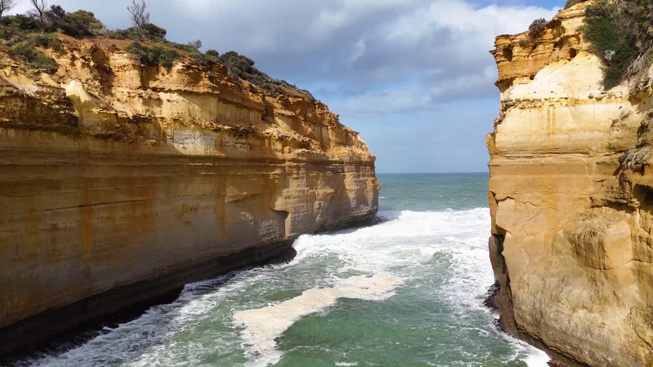Aerial footage of Loch Ard Gorge's dramatic cliffs and ocean waves under bright daylight, showcasing natural beauty and rugged landscapes