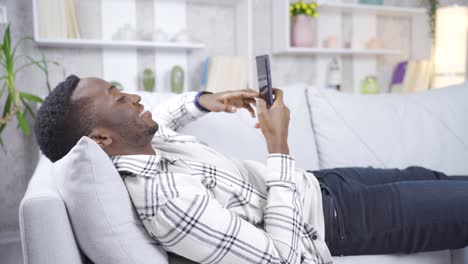 Glücklicher-Afrikanischer-Junger-Mann,-Der-Zu-Hause-Auf-Dem-Sofa-Liegt-Und-Lächelnd-Auf-Sein-Smartphone-Schaut