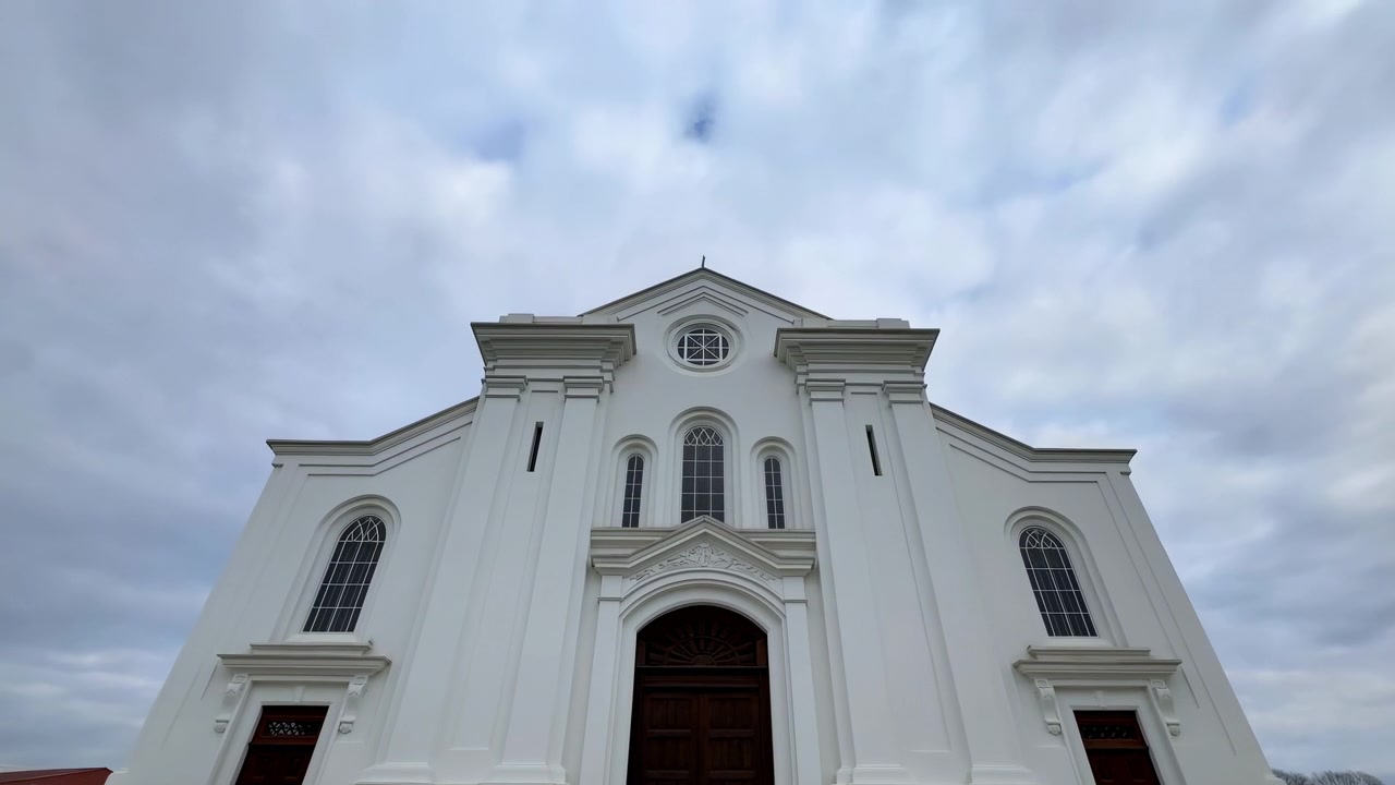 Vista desde un ángulo bajo que muestra la fachada de una iglesia clásica blanca con columnas simétricas, entrada arqueada, molduras decorativas y detalles desgastados bajo un amplio cielo nublado, creando una presencia monumental y serena