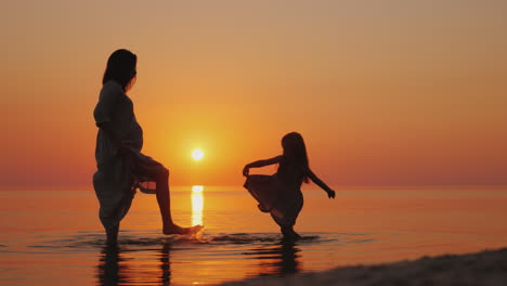 pregnant woman with her daughter having fun on the beach splashing water on each other waiting for t