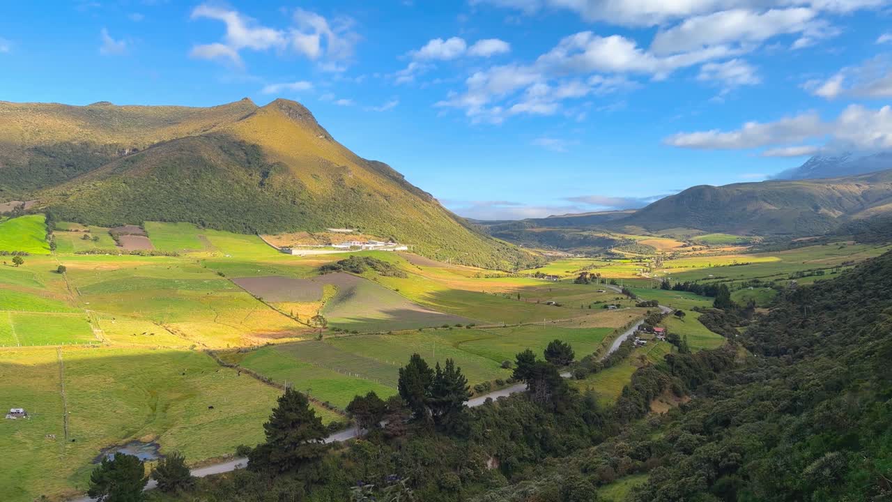 Ripresa a mano di una valle verde e pittoresca nelle Ande con una strada tortuosa, aziende agricole rurali e cime montuose sotto un cielo blu in Ecuador