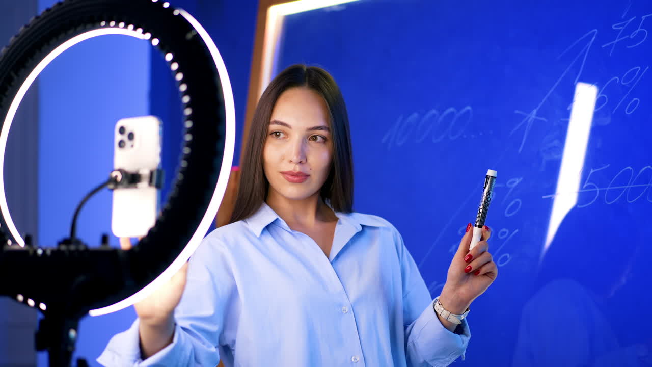 Beautiful Caucasian woman stands at the phone with light ring set for footage. Blogger turns to glass wall to write numbers on it.