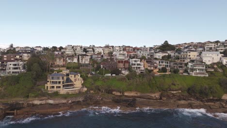 aerial drone slide right over sydney oceanfront mansion – cliffside waves, blue water, luxury rooflines, coastal neighborhood, morning sky in nsw