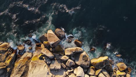 Cinematic-drone-shot-in-50Fps-flying-upwards-looking-down-at-rocky-coastline-with-dark-ocean-waves-crashing-against-the-boulders