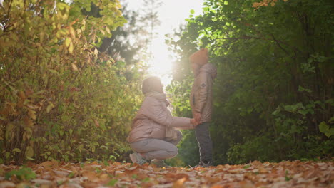 caring mother kneels in autumn woods, straightening daughter hoodie under warm sun halo, both in jackets and hat amidst colourful foliage, sharing joyful tender moment of love and connection