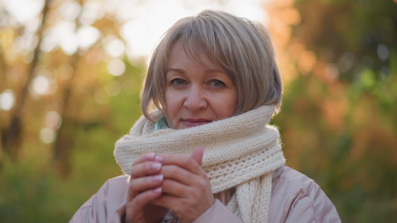 elegant mature woman seated outdoors in golden autumn light, wrapped in knit scarf, cupping hands around warm mug as sunlight halo glows around head, expression serene and thoughtful