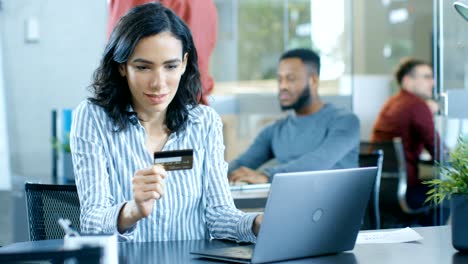 beautiful female office worker makes internet purchase from her laptop using credit card. in the background creative young people working.