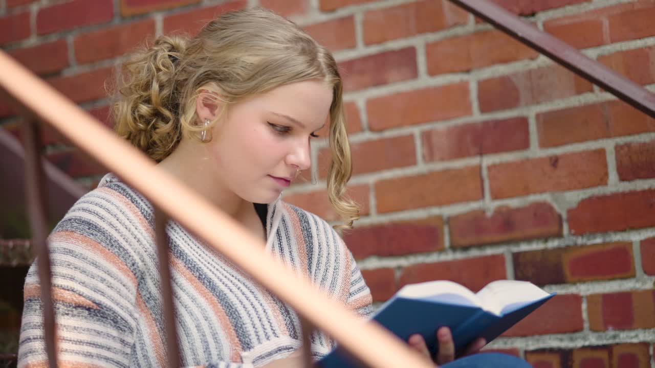 Studentessa legge un libro e volta le pagine, seduta all'aperto sulle scale accanto a un muro di mattoni.
