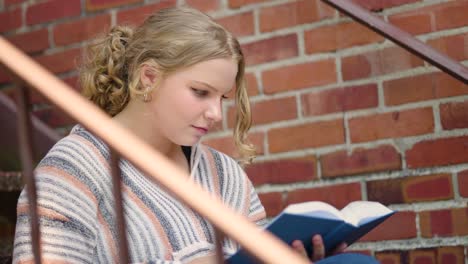 Girl-student-reads-book-and-turns-pages-sitting-outside-on-stairs-next-to-a-brick-wall