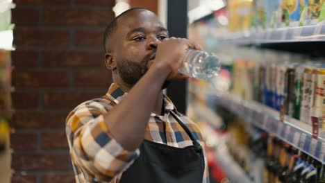 a man with black skin color in a plaid shirt drinks water from a transparent plastic bottle in a supermarket