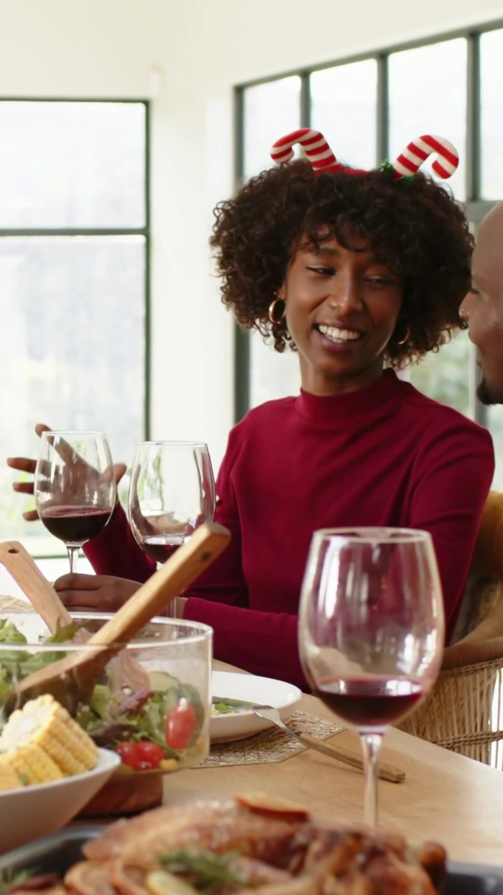 Vertical video: Smiling woman in red sweater holding wine, talking at holiday meal as man entering