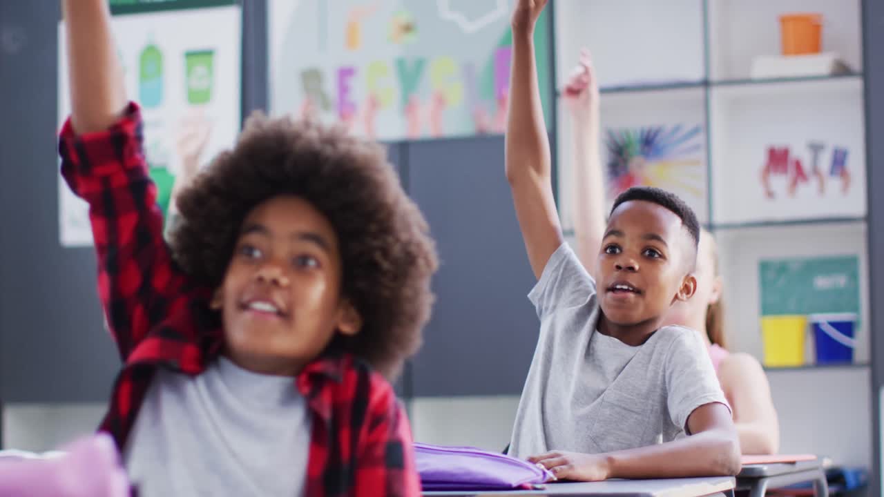 Happy diverse schoolchildren at desks raising hands in school classroom