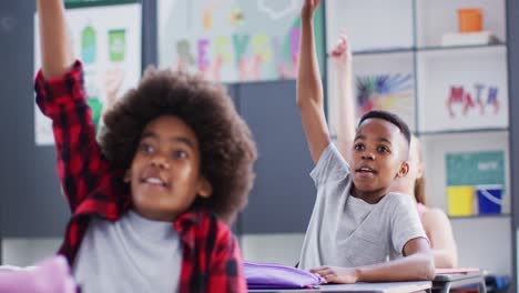 happy diverse schoolchildren at desks raising hands in school classroom