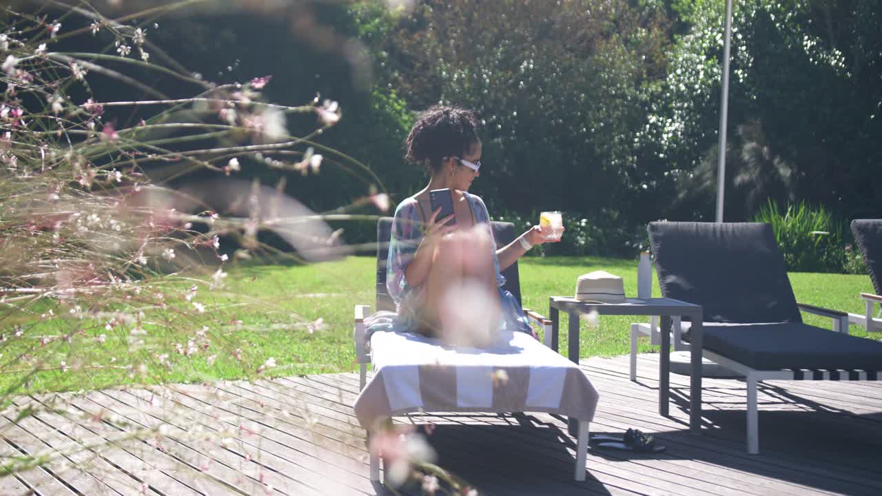 Adult woman wearing shades, sitting on lounge chair on deck, handling smartphone, raising glass