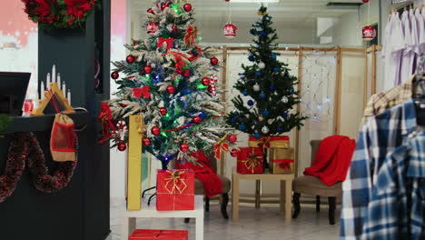 dolly in shot of festive clothing store filled with beautifully decorated christmas trees, garlands and ornaments during winter holiday season. empty fashion boutique with clothes on racks